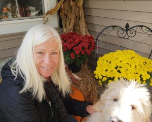 Kris Oleksyn on her front porch with her dog Charlie surrounded by fall decorations and red and yellow mums.